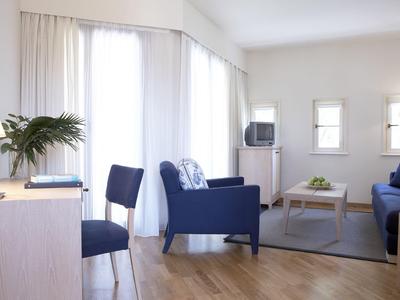 Helles Wohnzimmer mit blauen Sesseln, Couch, Holzfußboden, Tisch und großen Fenstern mit weißen Vorhängen.