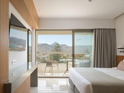 Hell eingerichtetes Hotelzimmer mit Doppelbett, Fernseher, Balkon und Bergblick bei klarem Himmel.