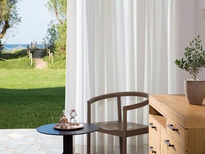 Moderner Holztisch und Stuhl vor weißen Vorhängen mit Blick auf einen Garten und das Meer.