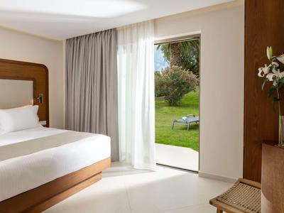 Helles Hotelzimmer mit großem Bett, Vorhängen und Blick auf grünen Garten.