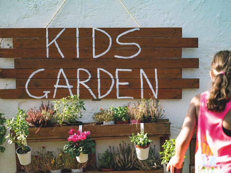 Een houten bord met de tekst 'Kids Garden' boven bloempotten met diverse planten, met een kind ervoor.
