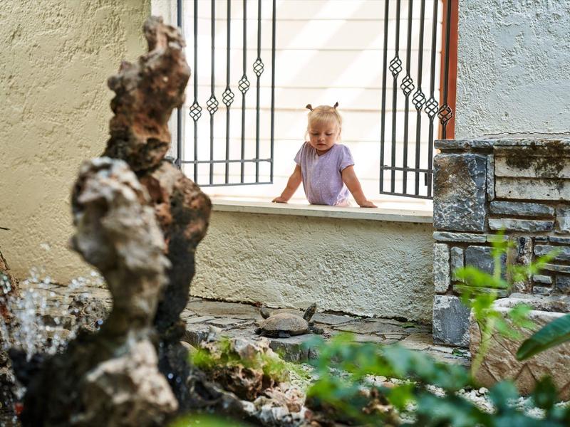 Een klein meisje leunt op een vensterbank en kijkt in een tuin met een sculptuur.