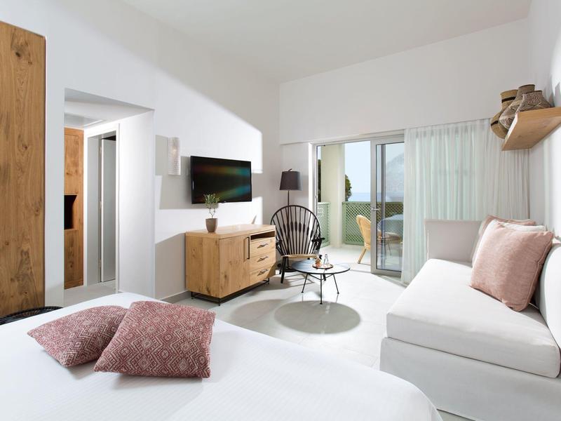 Licht hotelkamer met moderne inrichting, bank, bed, tv en toegang tot balkon.