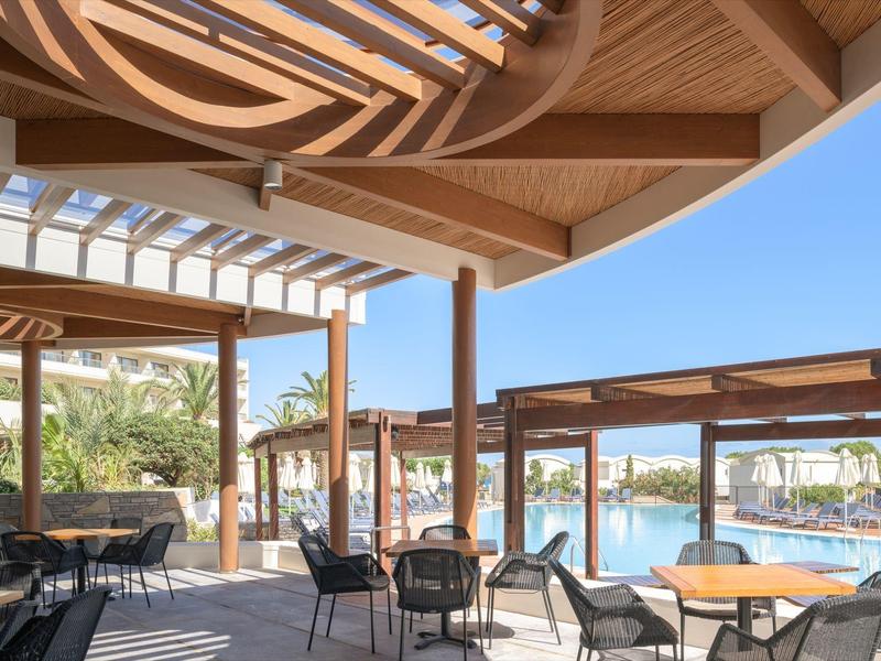 Overdekt terras met tafels en stoelen met uitzicht op een zwembad en ligstoelen bij heldere lucht.