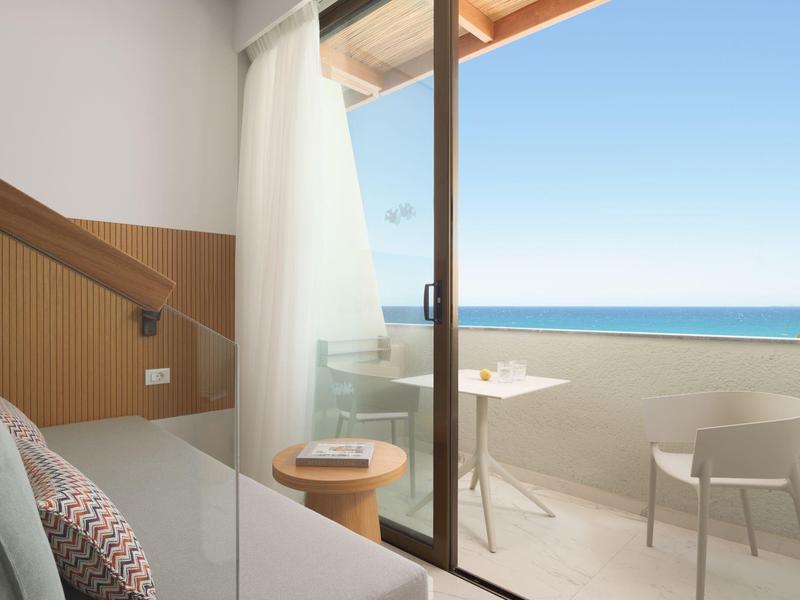 Modern hotelkamer met balkon, zeezicht en minimalistisch meubilair bij heldere hemel.