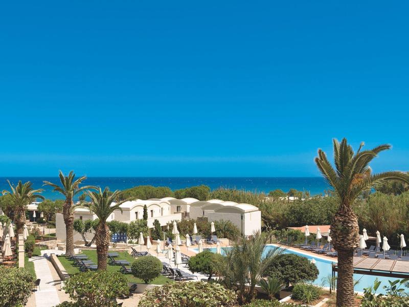 Uitzicht op een hotelzwembad met palmbomen en zee op de achtergrond onder een heldere blauwe lucht.