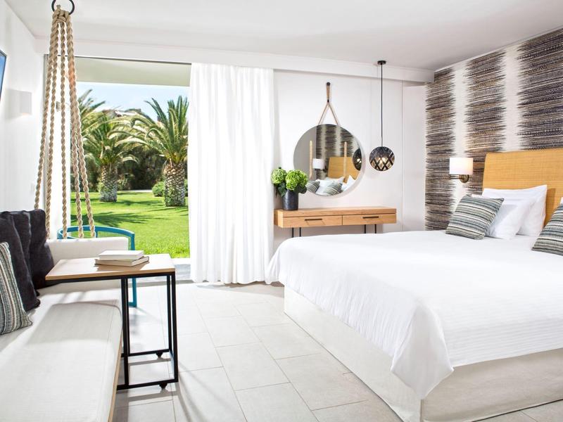 Lichte hotelkamer met tweepersoonsbed, zithoek en uitzicht op een tuin met palmbomen.