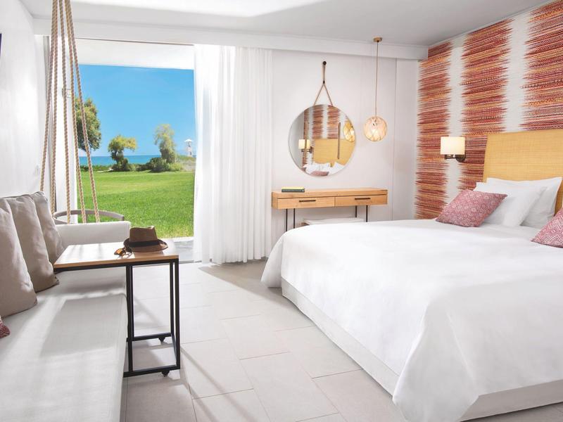 Licht ingericht hotelkamer met tweepersoonsbed, bank, raam met zeezicht en modern decor.