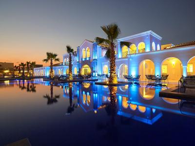 Hôtel éclairé avec piscine et palmiers au coucher du soleil se reflétant dans l'eau.
