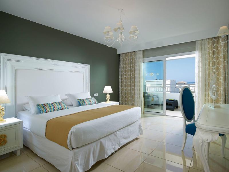 Lichte hotelkamer met kingsize bed, groene accentmuur, kroonluchter, balkon en stadszicht.