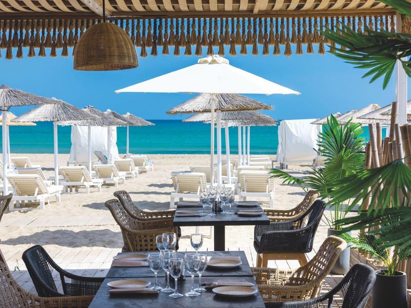 Eetgedeelte aan het strand met tafels, stoelen, parasols en uitzicht op zee en zandstrand.