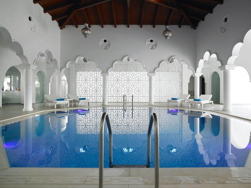 Binnenzwembad met bogenramen, witte muren en houten plafondbalken.