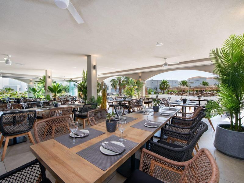 Openluchtrestaurant met moderne stoelen, houten tafels en planten met uitzicht op een mooie omgeving.