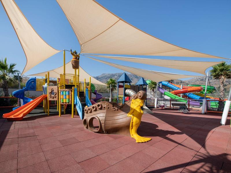 Speeltuin voor kinderen met glijbanen en schommels onder schaduwzeilen bij een zonnig resort.