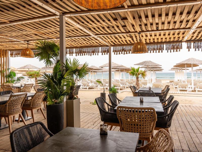 Overdekt strandterras met tafels, stoelen en uitzicht op zand en parasols.