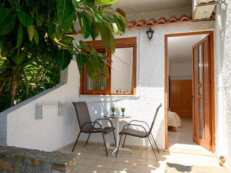 Pequeña terraza con mesa y dos sillas junto a la puerta de una habitación de hotel, rodeada de vegetación.