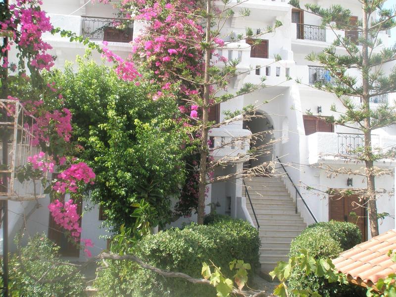 Edificio blanco brillante con escaleras, árboles y arbustos rosas florecientes en un jardín de hotel.
