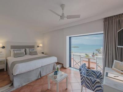 Helles Hotelzimmer mit Doppelbett, Sitzbereich und Meerblick durch ein großes Fenster.