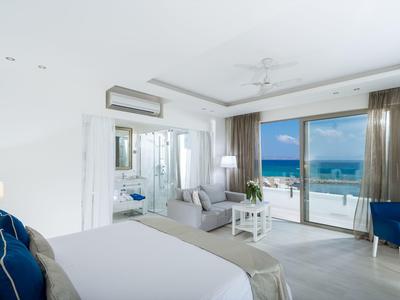 Helles Hotelzimmer mit Bett, Sesseln und Balkon mit Meerblick bei klarem Wetter.