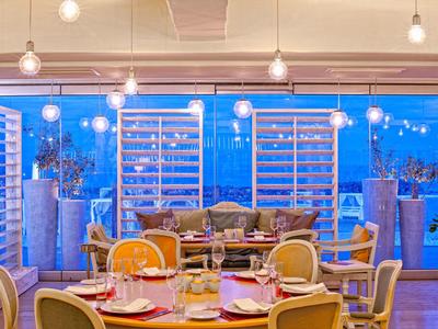 Restaurante elegante con mesas redondas, sillas y grandes ventanas que dan a un exterior iluminado de azul.