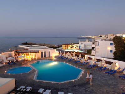 Vista notturna di una piscina esterna illuminata con lettini di fronte al mare vicino a edifici bianchi.