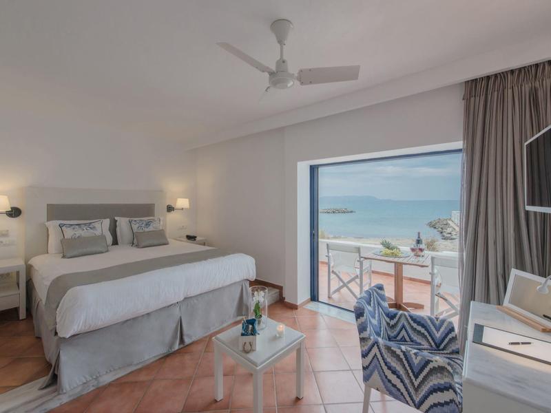 Helles Hotelzimmer mit Doppelbett, Sitzbereich und Meerblick durch ein großes Fenster.