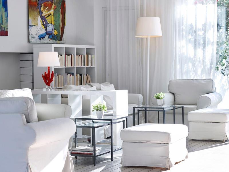 Helles Wohnzimmer mit weißen Möbeln, Teppich und großer Fensterfront, helle Vorhänge und Pflanzen.