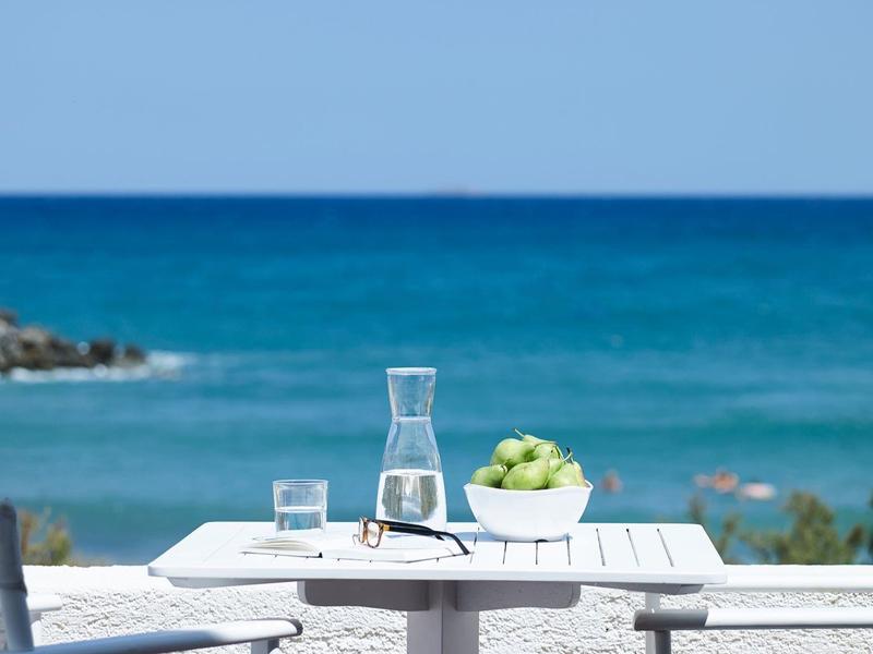 Weißer Gartentisch mit Karaffe, Gläsern und grünem Apfelschale vor blauem Meer und Himmel.