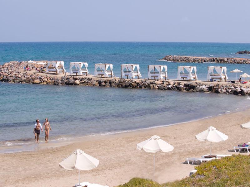 Leerer Sandstrand mit weißen Schirmen, zwei Personen am Wasser, blauer Himmel und ruhiges Meer.