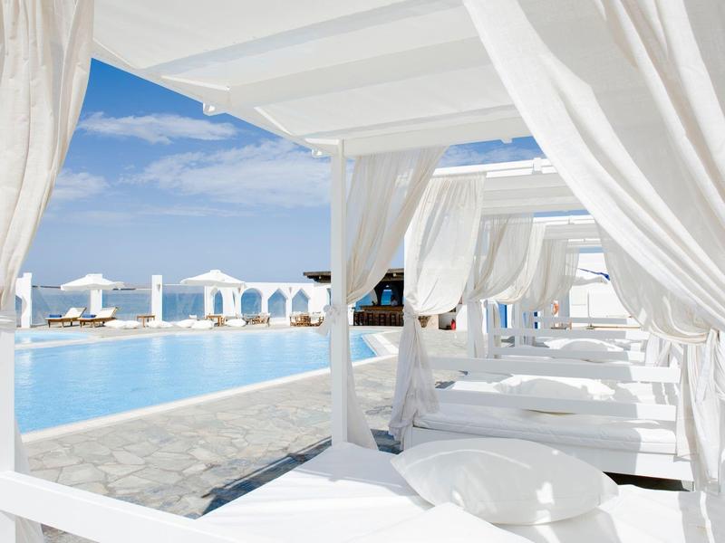 Weiße Sonnenliegen mit Vorhängen stehen am blauen Pool unter klarem Himmel am Meer.