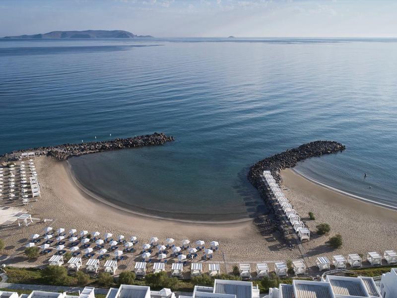 Strand mit Liegestühlen und Sonnenschirmen neben ruhigem Meer und mehreren Wellenbrechern.