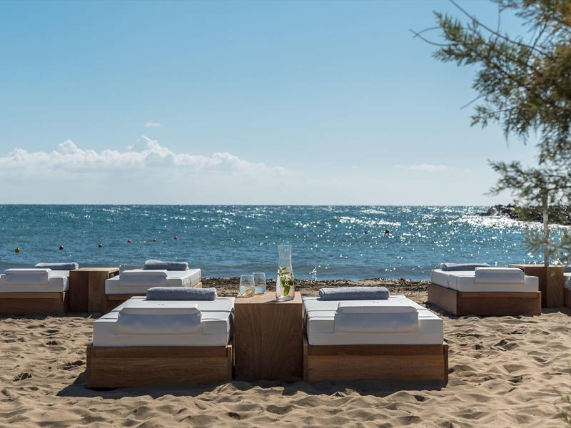 Strand mit Sonnenliegen aus Holz, kleinen Tischen und blauen Himmel über dem ruhigen Meer.