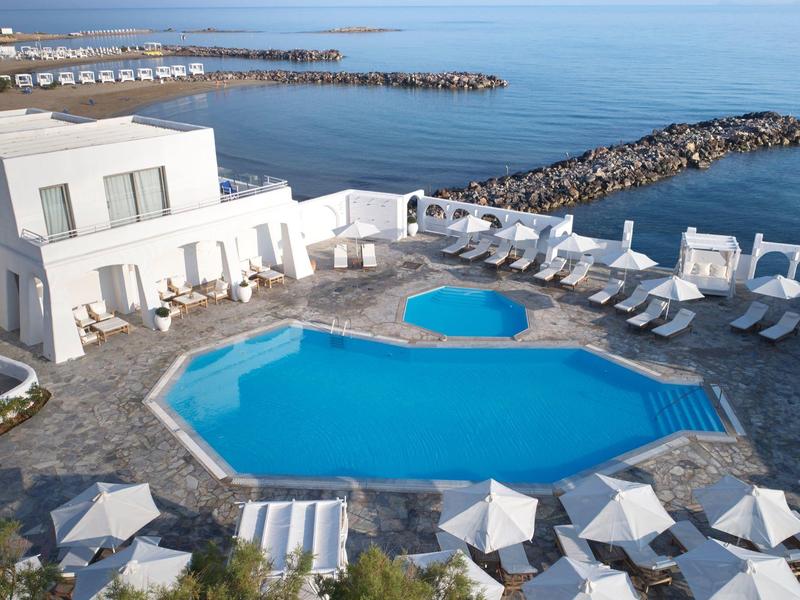 Ein großer blauer Pool umgeben von weißen Liegestühlen und Gebäuden am Meer mit Felsenwellenbrechern.