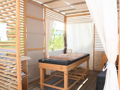 Luminoso spazio benessere con lettino da massaggio, rivestimenti in legno e tenda bianca.