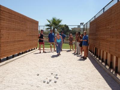 Persone che giocano a bocce su un campo di sabbia tra barriere di legno sotto un cielo azzurro.