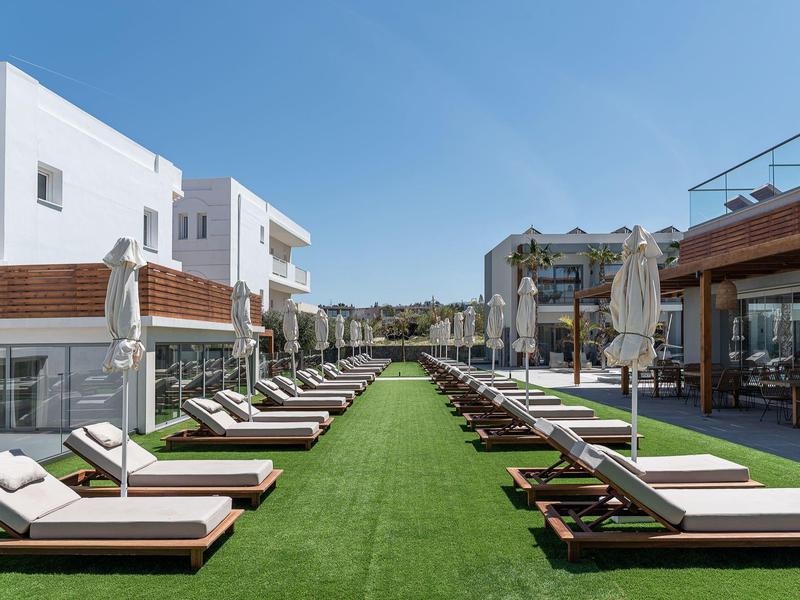 Due file di lettini con cuscini su prato verde tra edifici bianchi moderni sotto un cielo limpido.