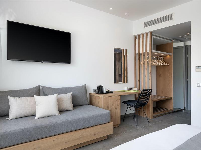 Camera d'hotel moderna con divano grigio, TV a parete e guardaroba aperto in legno