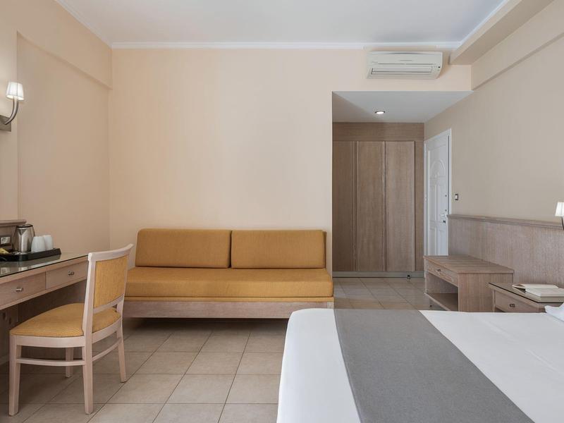 Camera d'albergo con letto, divano, scrivania e cassettiera in tonalità neutre.