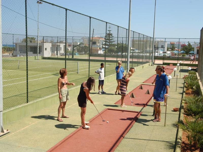 Diverse persone giocano a minigolf su un campo sportivo soleggiato accanto a un campo da tennis.