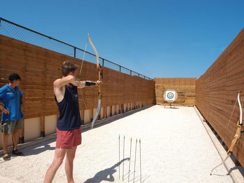 Due uomini che praticano il tiro con l'arco su una corsia sabbiosa con un bersaglio, recinzioni di legno e cielo azzurro.