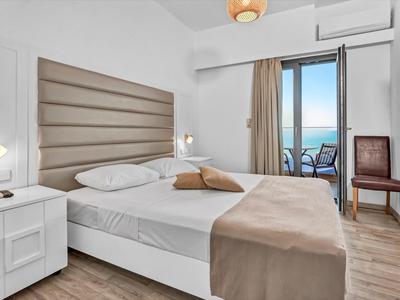 Helles Hotelzimmer mit Doppelbett, Nachttisch, Fernseher und Blick auf Balkon mit Meerblick.