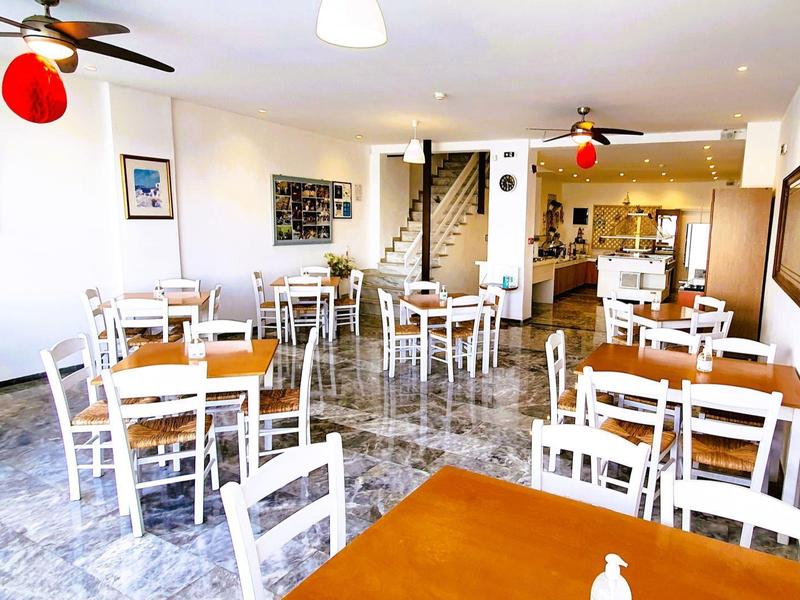 Helles, geräumiges Café mit weißen Holzmöbeln, grauem Boden und offenen Zugang zum Küchenbereich.