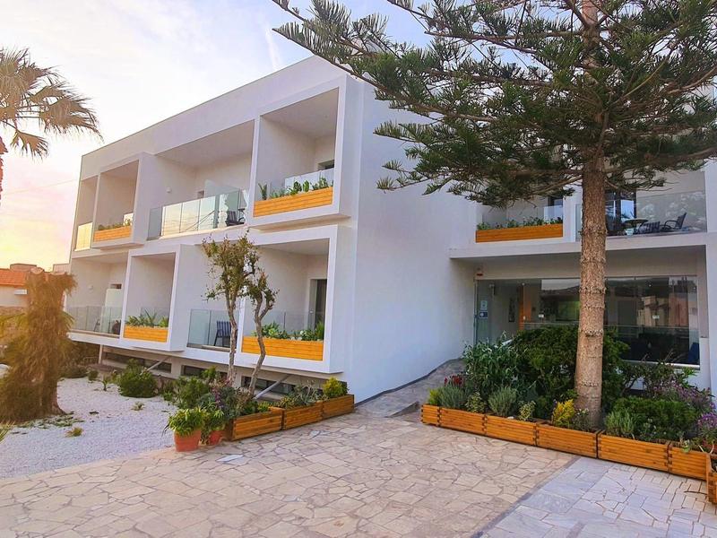 Modernes, weißes Gebäude mit Balkonen, Palmen und Bäumen, Pflanzbeete und gepflasterter Weg bei Sonnenuntergang.