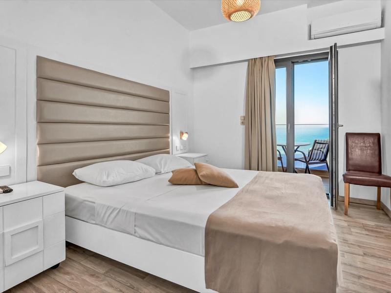 Helles Hotelzimmer mit Doppelbett, Nachttisch, Fernseher und Blick auf Balkon mit Meerblick.