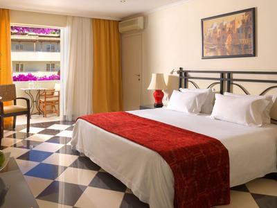 Helles Hotelzimmer mit Doppelbett, roten Details, Stühlen und Balkon mit Blick nach draußen.