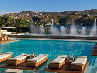 Outdoor-Pool mit Liegestühlen und Wasserspielen vor bergiger Landschaft bei klarem Himmel.