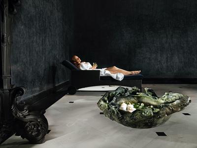 Osoba odpoczywa na leżaku w ciemnym pokoju z dekoracyjnym zielonym obiektem na podłodze.