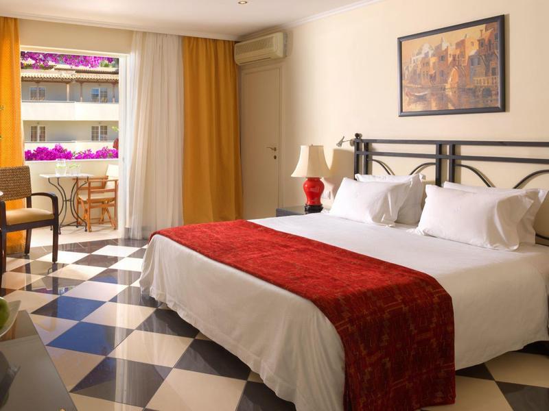 Jasny pokój hotelowy z dużym łóżkiem, czerwonymi akcentami i balkonem z roślinami.