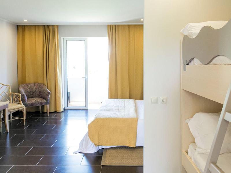 Jasny pokój z łóżkiem podwójnym, łóżkiem piętrowym, fotelem i żółtymi zasłonami przy drzwiach na balkon.