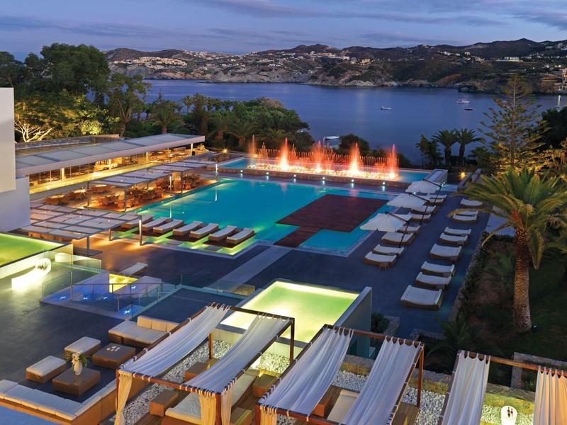 Strefa basenowa hotelu z paleniskami i leżakami, widok na jezioro i góry o zmierzchu.